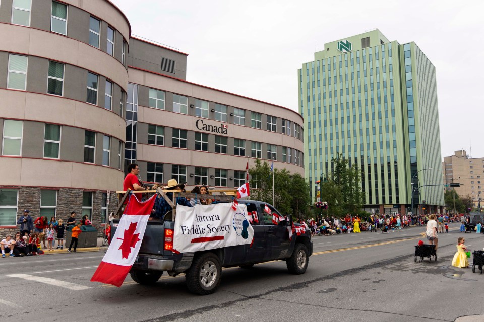 The Aurora Fiddle Society float passes the federal Greenstone Building (notably not the greenest building in the photo).