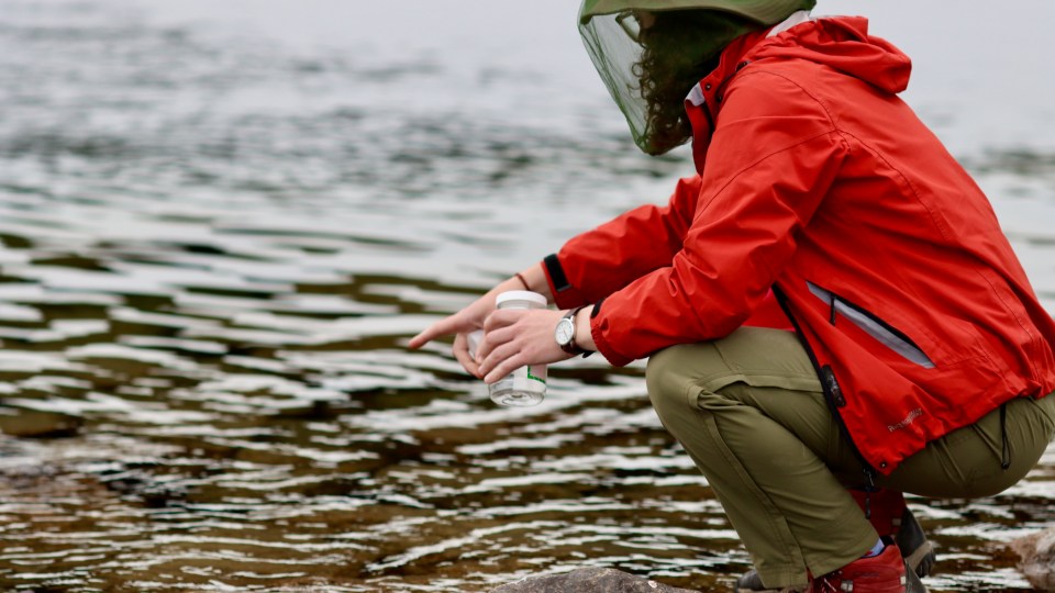 Baran collects a water sample from the La Martre River. Chloe Williams/Cabin Radio