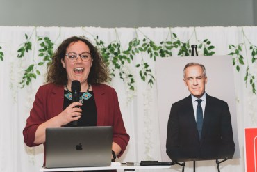Liberal candidate Rebecca Alty speaks in front of a Mark Carney poster. Sarah Pruys/Cabin Radio