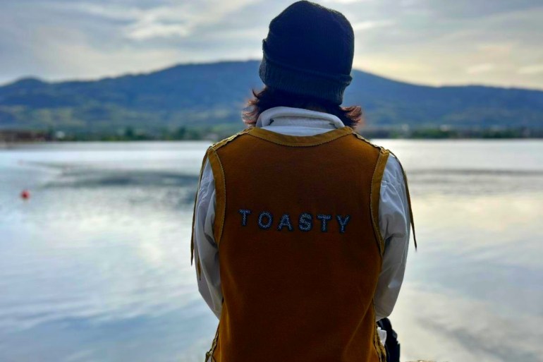 Chief of West Point First Nation, Devon Felker sits with his back facing the camera. He wears a brown vest with the word 'Toasty' across his back. He is onlooking a waterfront view with mountains seen in the background.