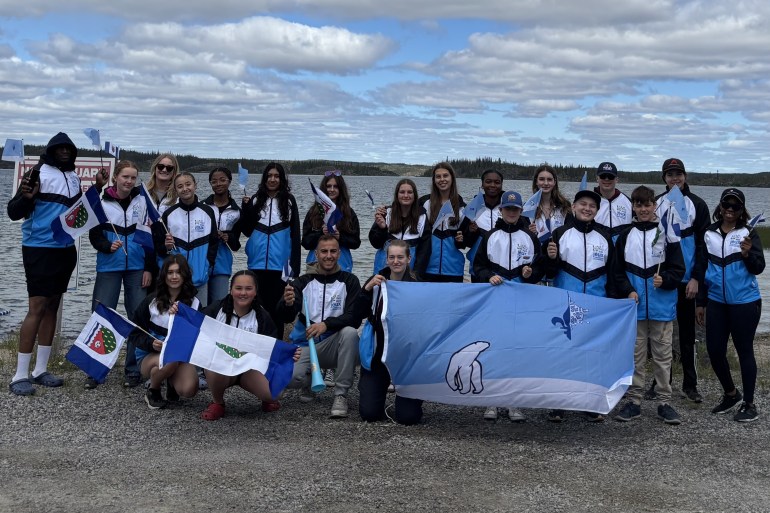 Group of NWT youth wearing white and light blue jackets pose holding NWT flags.