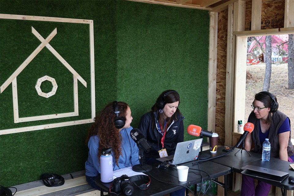 3 women seated at a table talking into microphones. They sit in front of an image of a cabin.