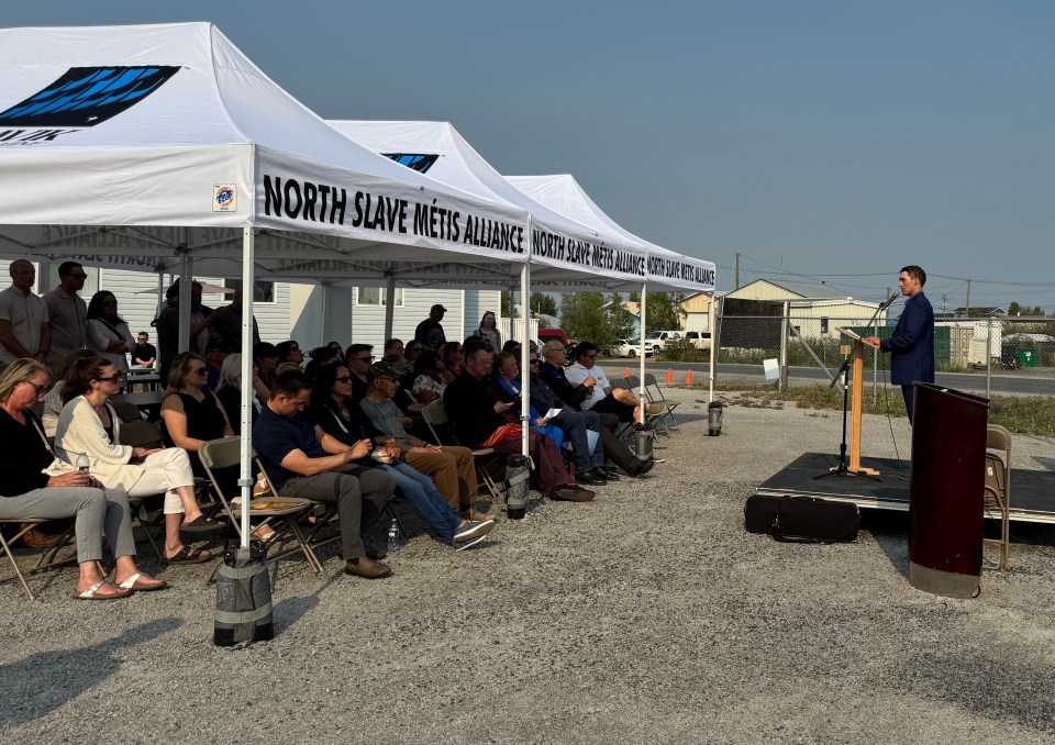 A crowd sits under a white canopy reading North Slave Métis Alliance on the canvas. A speaker stands facing them speaking into the mic.