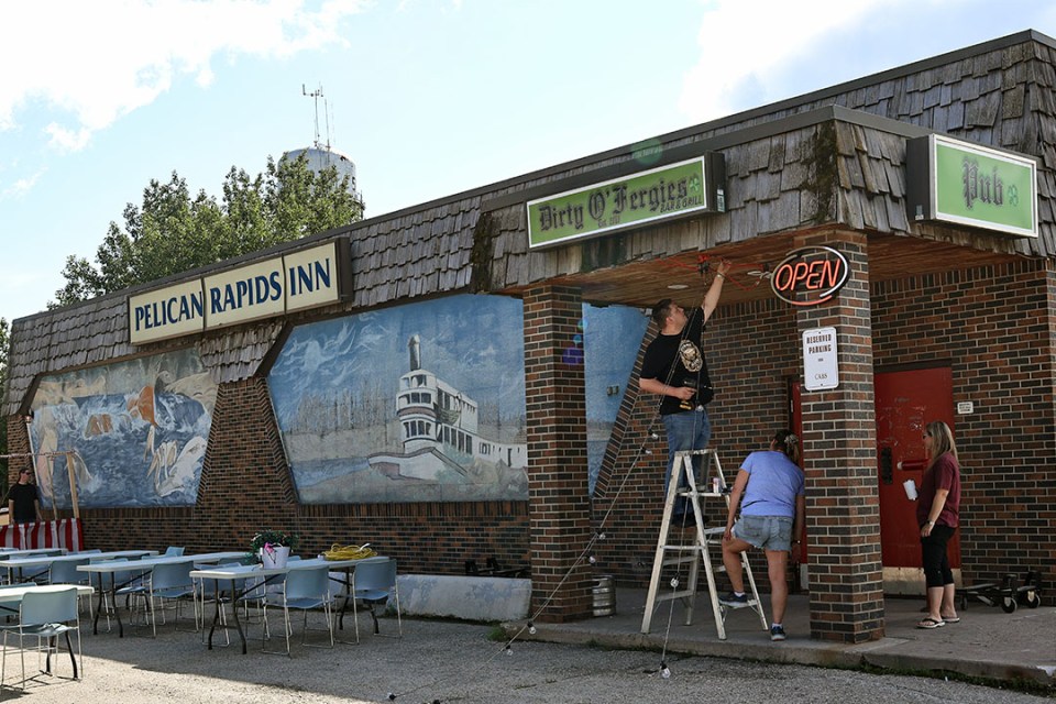 A woman holds a ladder steady for a man at the top attaching lights to a building.