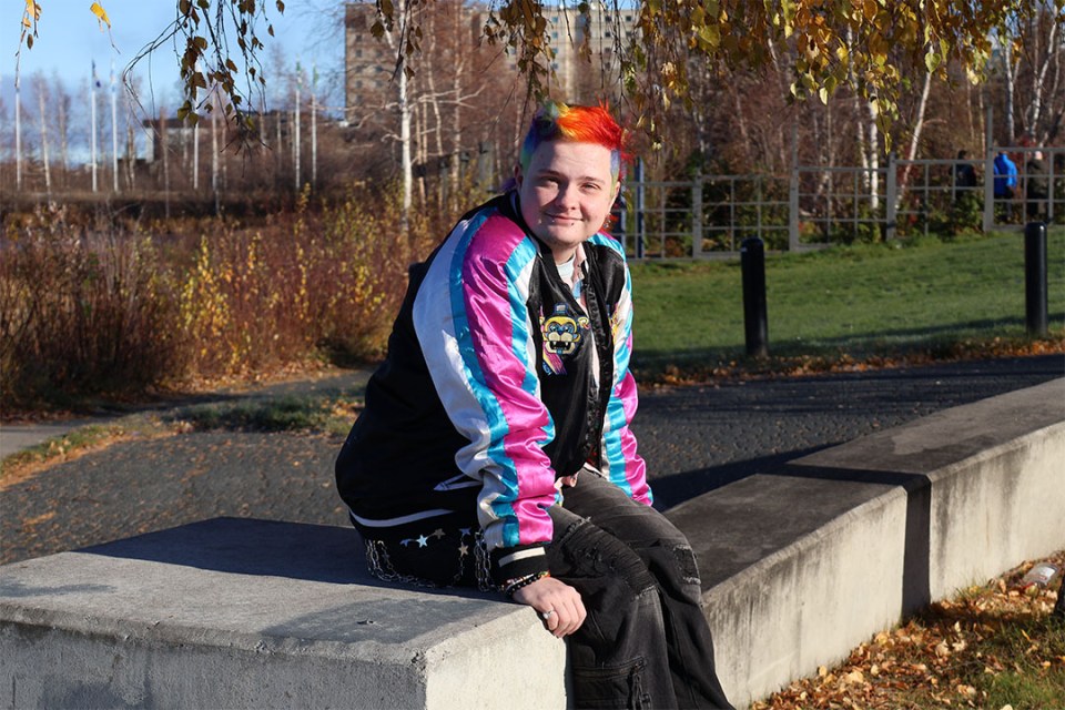 Person with rainbow hair sitting on a concrete barrier in the sunlight.
