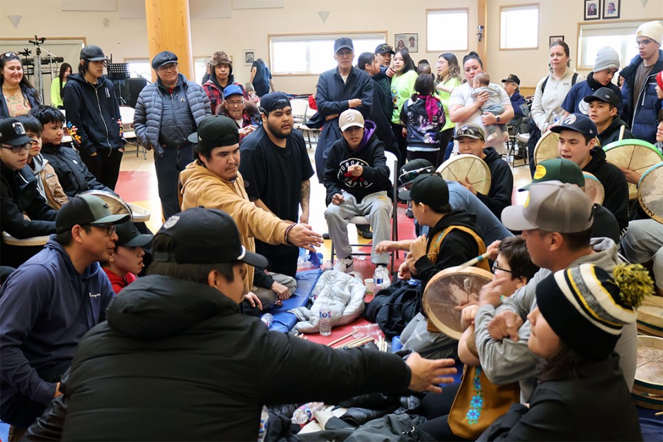 A group of men and boys play hand games, with onlookers watching 