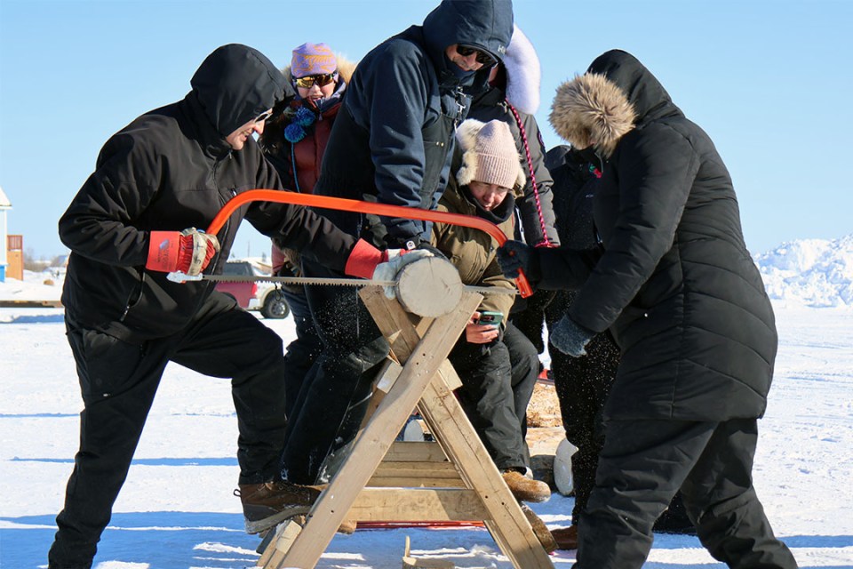 Two people saw through a log as part of a game. Several people sit on the log to help keep it steady