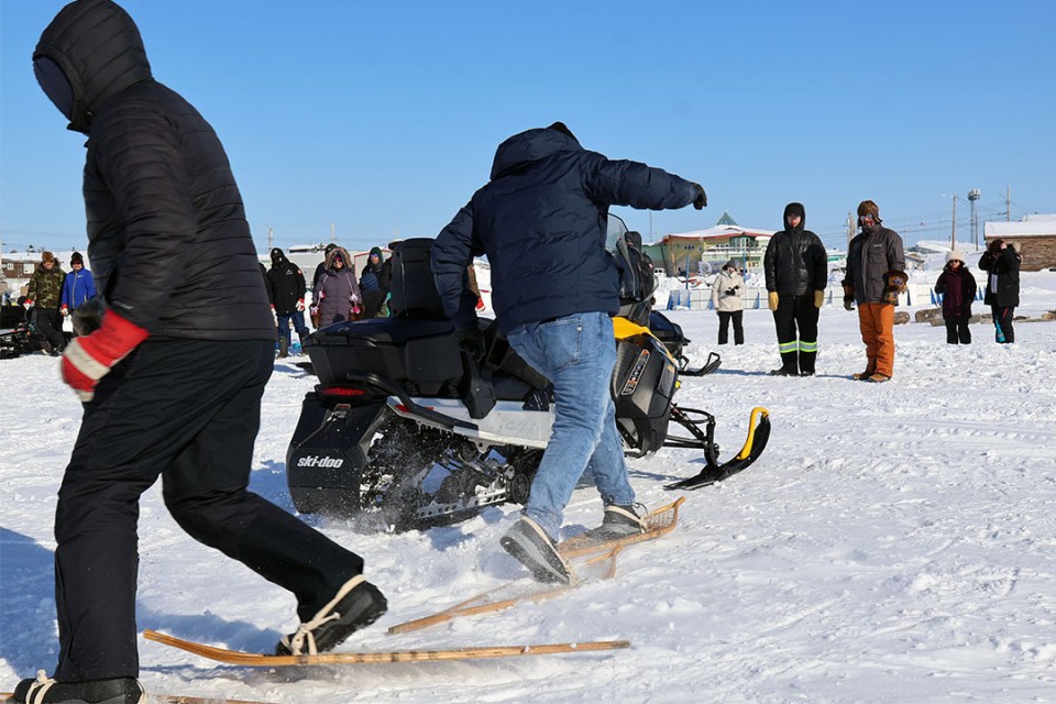 Two people wearing snowshoes race around a skidoo