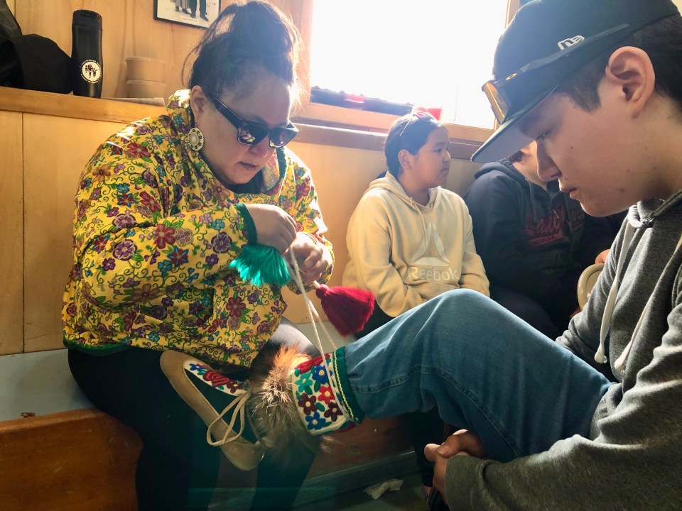 Diandra Raddi-Kuiksak helps her son Trent Wolki do up the kamiks that belonged to his grandfather ahead of the drum dance. Cody Punter/Cabin Radio